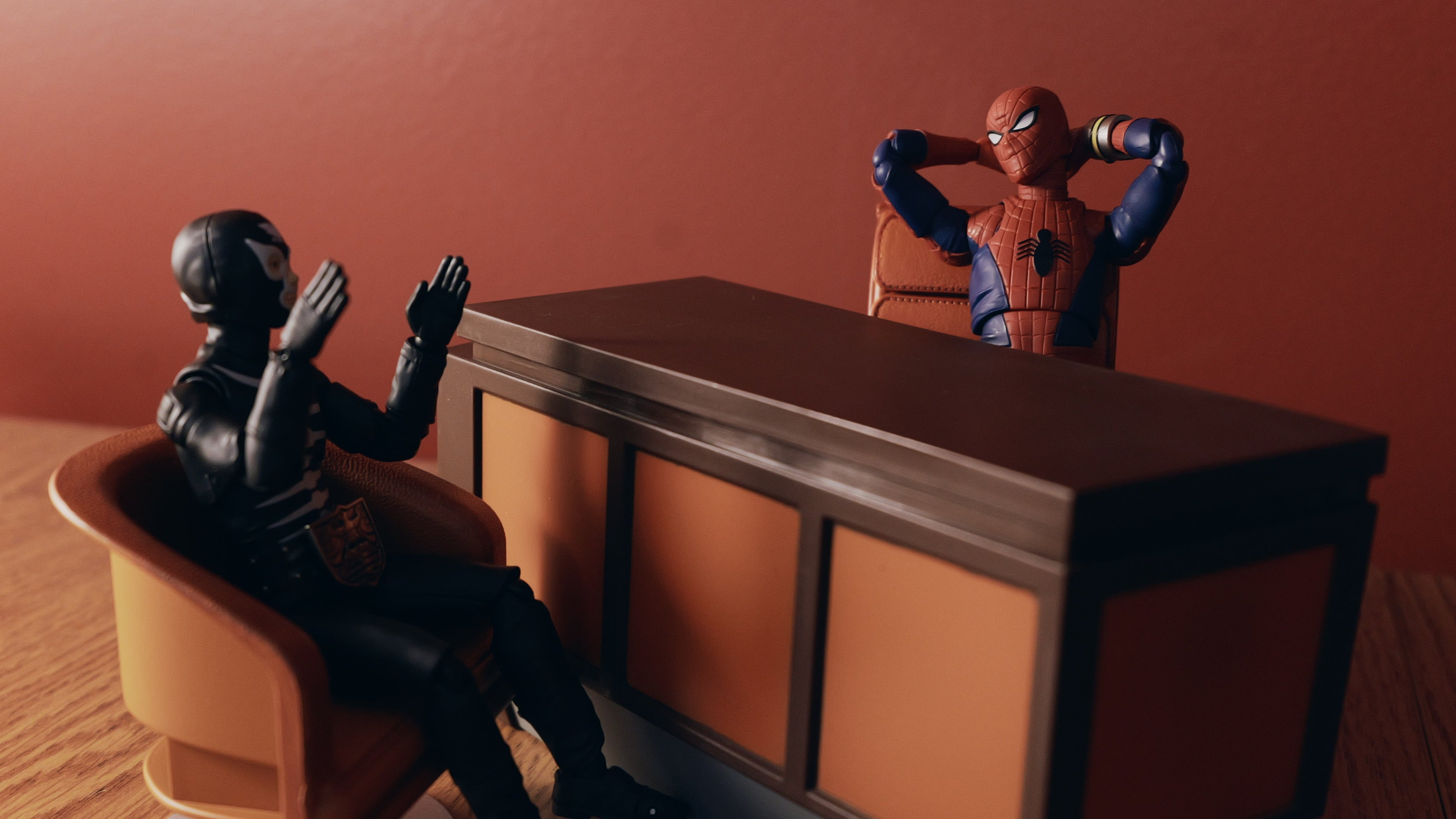 Two superhero figurines sitting at a desk against a red wall.
